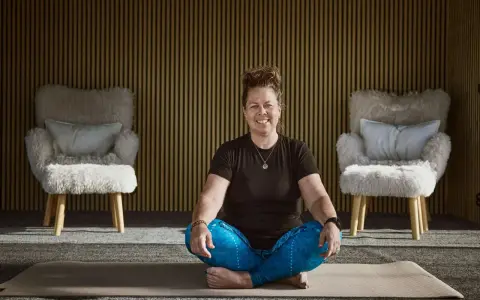 Kvinna i meditationsställning framför en vägg med träribbor. Två fluffiga fåtöljer står på var sin sida. Kanske hon funderar på hur hon ska dekorera hylla i sitt hem.