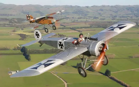 Zwei historische Fokker Eindecker fliegen über grüne Felder. Ein Pilot im Vordergrund, der andere im Hintergrund.