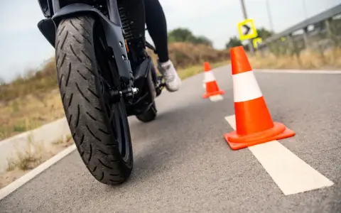 Motociclista practica maniobras entre conos naranjas. La rueda delantera de la moto, con su banda de rodadura, se ve de cerca.