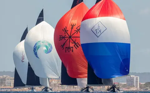 Segelboote der j class rasen über das Meer. Bunte Spinnaker mit Logos und Symbolen, im Hintergrund eine Küstenstadt.