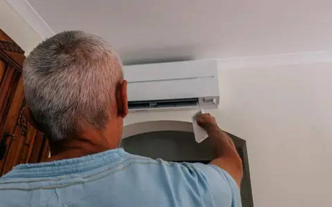 Uomo con telecomando regola il condizionatore per aria fredda, cercando sollievo dall'estate.