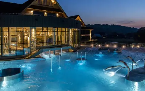Nocą w Termach Chochołowskich hotel lśni światłem, a baseny zewnętrzne kuszą błękitem. Ludzie pływają i relaksują się w wodzie.