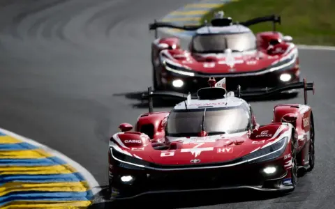 Dos coches de carreras rojos compiten en Le Mans, sus luces brillan en la pista.