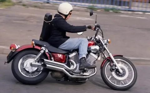 Mężczyzna w kasku jedzie na czerwonym motocyklu Yamaha Virago 250 z futerałem na plecach.