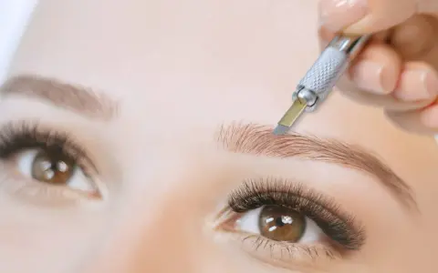Main d'une esthéticienne réalisant un **tatouage sourcils poil par poil** avec un outil fin. Gros plan sur un œil et un sourcil.