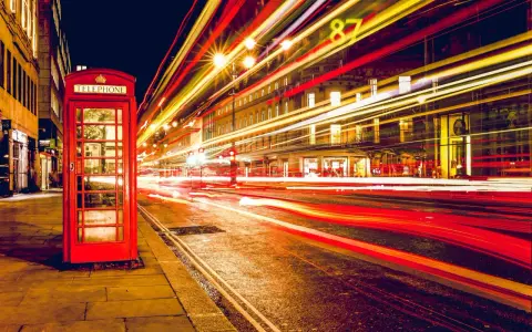 Klasyczna czerwona budka telefoniczna na tle nocnej ulicy, gdzie długi czas naswietlania zdjecia uchwycił smugi świateł samochodowych.