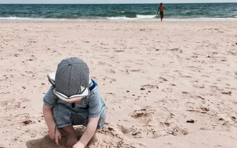 Kleines Kind mit Hut spielt im Sand am Strand der Algarve. Im Hintergrund läuft ein Mann ins Meer. Ein perfekter Tag für Algarve mit Kindern.