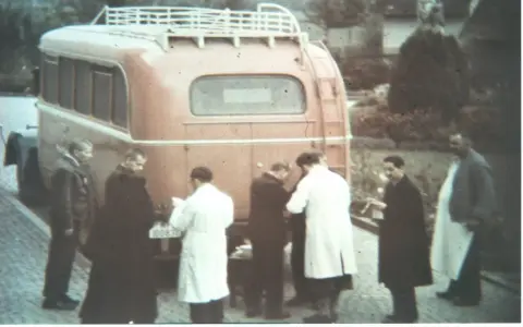 Männer in weißen Kitteln und dunklen Mänteln stehen vor einem roten Bus. Ein Kennzeichen ist sichtbar. Die Szene könnte mit der NS-Euthanasie in Verbindung stehen.