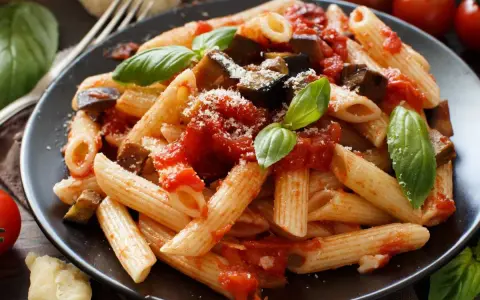 Un plato de pasta alla norma con penne, berenjena, salsa de tomate y albahaca fresca, espolvoreado con queso rallado.