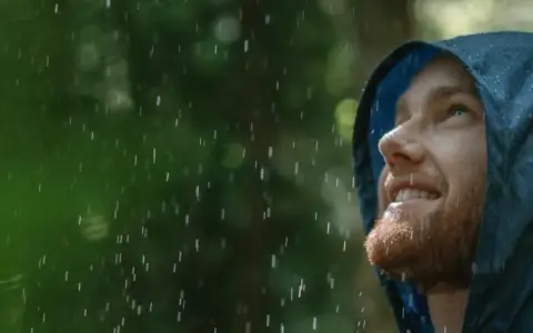 Mann mit Bart blickt lächelnd in den Regen, seine blaue Regenjacke hält ihn trocken.