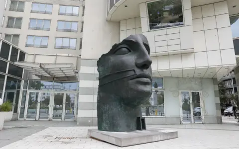 Okaleczona, ciemna rzeźba głowy z opaską na ustach, na tle nowoczesnego budynku.
