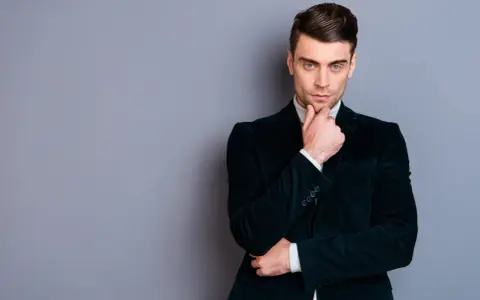 Elegancki mężczyzna w ciemnej, **marynarce aksamitnej męskiej**, z dłonią na podbródku, spogląda wprost.