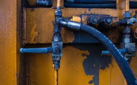 Détail d'un système hydraulique avec un tuyau noir marqué "HANSA-FLEX HD 24", un levier bleu et un sertissage flexible sur fond jaune usé.
