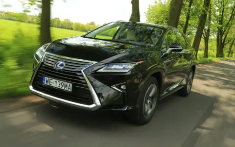 Czarny Lexus RX 450h pędzi po wiejskiej drodze, otoczony zielenią drzew i pól.