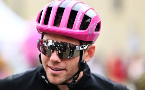 Cyclist Luke Lamperti smiles, wearing a bright pink helmet and reflective sunglasses.