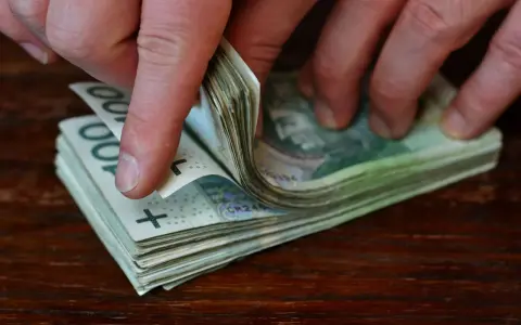 Dłonie przewracają stos banknotów 100 zł na drewnianym blacie.