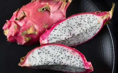 Cały i przekrojony smoczy owoc na czarnym talerzu. Biały miąższ z czarnymi pestkami kusi, by odkryć, jak smakuje smoczy owoc.