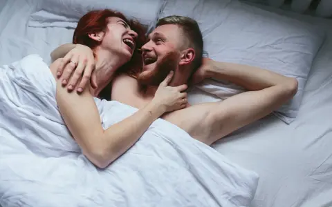 Para kochająca się w łóżku, śmiejąca się. Kobieta z rudymi włosami obejmuje mężczyznę z brodą. Sikanie podczas seksu może być częścią intymnych chwil.