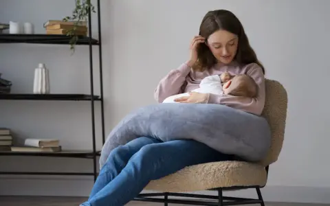 Szukasz idealny fotel do karmienia? Młoda mama z dzieckiem na rękach, otulona poduszką, siedzi na wygodnym, pluszowym fotelu.