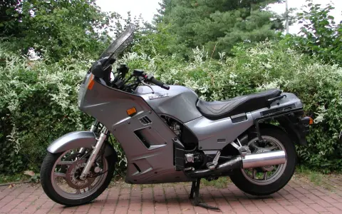 Srebrny motocykl Kawasaki GTR 1000 zaparkowany na czerwonej kostce brukowej, otoczony zielenią.
