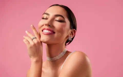 Mujer sonríe con ojos cerrados, luciendo un maquillaje romántico, joyas brillantes y un fondo rosa.