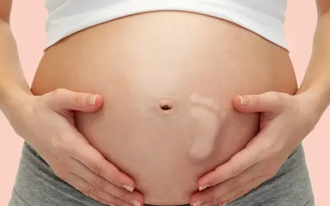 Brzuszek w 30 tygodniu ciąży, z widocznym odciskiem stópki dziecka. Ręce mamy delikatnie obejmują rosnący brzuch.