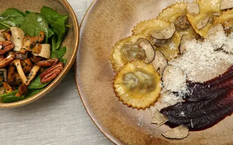 Ravioli pasta mit Trüffeln, Parmesan und einer dunklen Sauce. Daneben eine Schüssel mit Pilzen, Nüssen und Spinat.