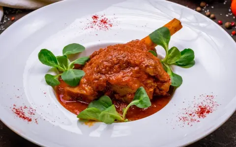 Pierna de cordero en salsa roja con hojas verdes en plato blanco.
