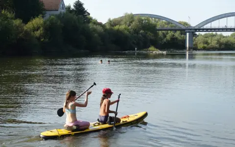 Zwei Mädchen genießen das Stand Up Paddling in Heidelberg. Im Hintergrund eine Brücke und Bäume.