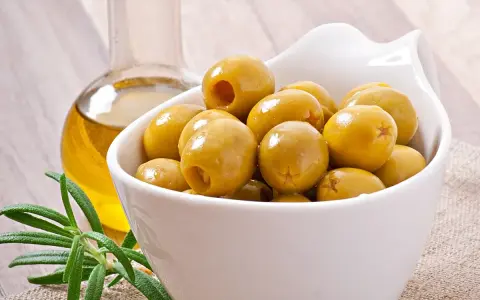 Aceitunas verdes en un cuenco blanco junto a una botella de aceite de oliva y romero. Descubre los aceitunas beneficios y contraindicaciones.
