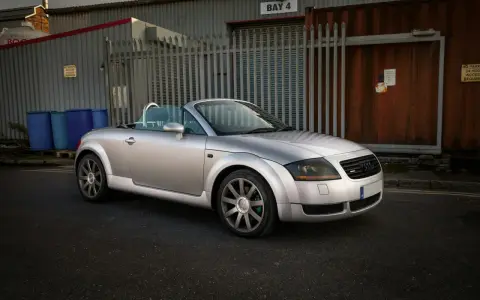Silberner Audi TT 2002 Roadster mit offenem Verdeck steht auf einer Straße vor einem Industriegebäude.