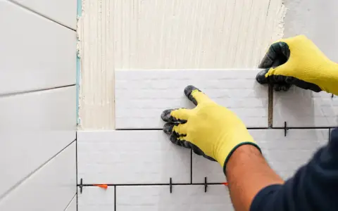 Mains gantées de jaune en train de coller du carrelage sur du carrelage, avec des croisillons pour un espacement parfait.