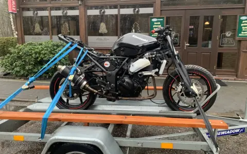 Czarny motocykl Kawasaki na przyczepce, zabezpieczony pasami. W tle budynek z napisem "Leśny Staw". Przepisy dotyczące przyczepki do motocykla są kluczowe dla bezpiecznego transportu.