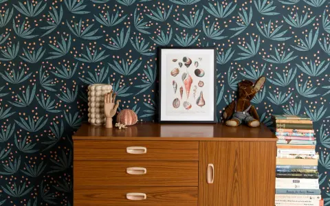 Idée motif peinture mur : papier peint botanique bleu nuit avec des feuilles stylisées et des baies roses. Un cadre de coquillages et une commode vintage complètent la scène.