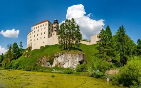 Zamek na wzgórzu, otoczony zielenią, z jeziorem w dole. Prawdziwa polska Szwajcaria, zapraszająca do podziwiania krajobrazów.