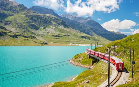 A red train winds along a track beside a vibrant turquoise lake, with lush green mountains and a bright blue sky in the background. This is a classic como to switzerland train journey.