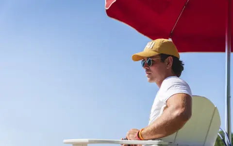 Ein Rettungsschwimmer mit gelber Kappe und Sonnenbrille wacht am Pool des **summer camp**.