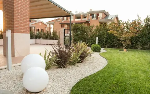 Aiuole davanti casa con ghiaia bianca, sfere luminose e prato verde curato. Sullo sfondo, case moderne e siepi.