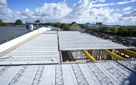 Budowa dachu kopertowego z elementami kratownicowymi i betonem. W tle budynki i błękitne niebo z chmurami.
