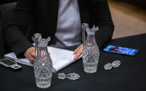 Une personne écrit sur un carnet, entourée de flacons en cristal. Une folle enchère se prépare pour ces objets précieux.