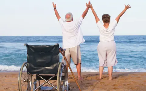 Szczęśliwi seniorzy cieszą się urlopem. Wózek inwalidzki i laska na plaży świadczą o tym, że to udany turnus rehabilitacyjny nad morzem.