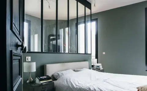 Agencement chambre moderne avec lit blanc, table de chevet industrielle et verrière séparant l'espace.