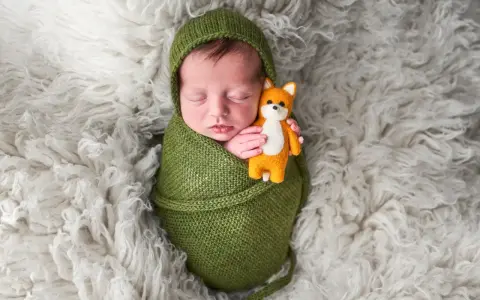 Dolci immagini neonati: un bimbo avvolto in una coperta verde tiene stretto un piccolo volpino di peluche, dormendo sereno su un soffice tappeto bianco.