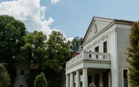 Pałac Grąbkowo, biały budynek z kolumnami i balkonem, otoczony bujną zielenią drzew pod błękitnym niebem z chmurami.