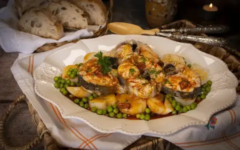 Merluza a la gallega servida en plato blanco con patatas, guisantes y perejil. Pan rústico en cesta al fondo.