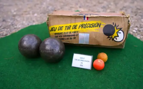 Deux boules de pétanque et un cochonnet sur un tapis vert. La boîte du jeu de tir de précision est visible. Apprenez comment tirer à la pétanque !
