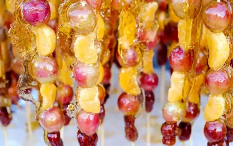 Spiedini di frutta caramellata con acqua e zucchero, con uva e spicchi d'arancia lucidi e dorati.