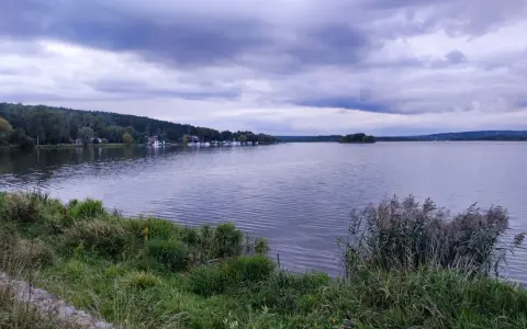 Spokojny zalew wióry, z łodziami przy brzegu i lasem w tle, pod burzliwym niebem.