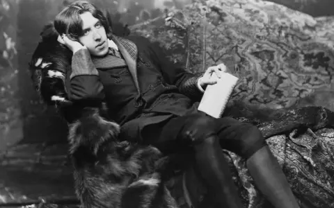 Oscar Wilde, w futrzanym płaszczu, z książką w dłoni, w pozie sugerującej refleksję nad swoimi sztukami.