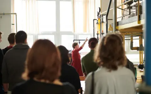 Grupa studentów na zajęciach praktycznych. Kobieta w czerwonej sukience tłumaczy coś przy aparaturze. To może być część ich studiów magisterskich zaocznych w Warszawie.